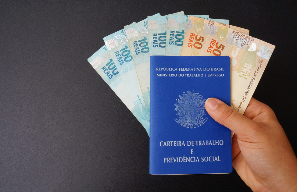 Woman's hand holding Brazilian Work Card (Carteira de Trabalho) and Brazilian money on black background. Work concept.